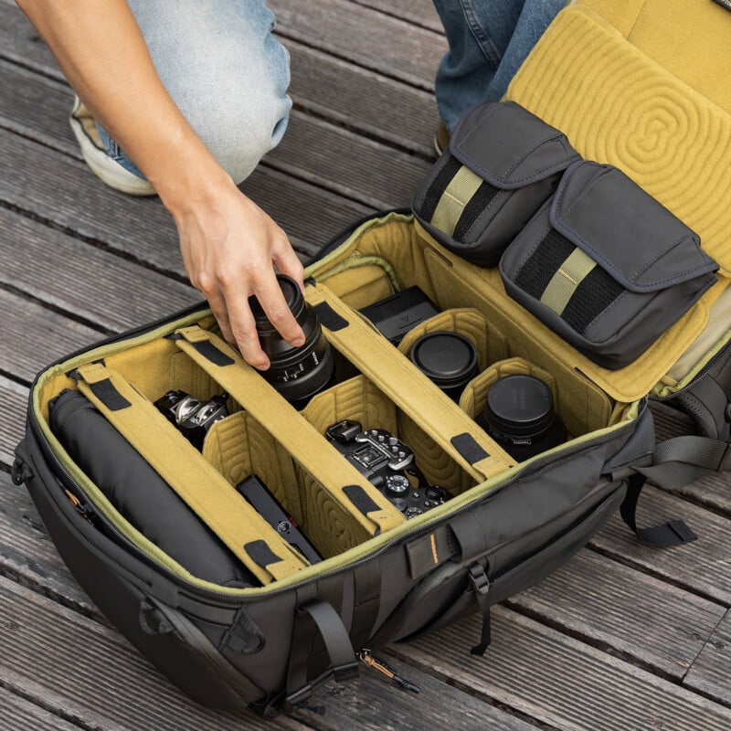 A person kneels on wooden planks, organizing a black camera backpack with neatly arranged camera bodies, lenses, and accessories in padded compartments.