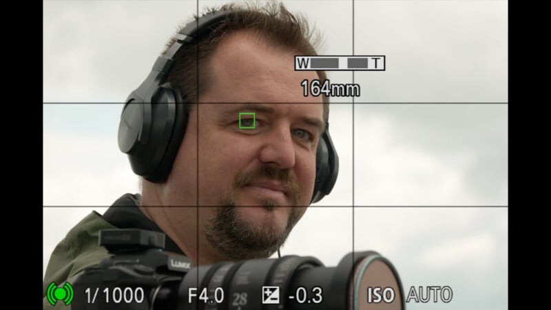 A man with headphones is seen through a camera viewfinder. Camera settings and focus grid are displayed on screen, with the focus point on his eye. He is holding a camera outdoors under a cloudy sky.