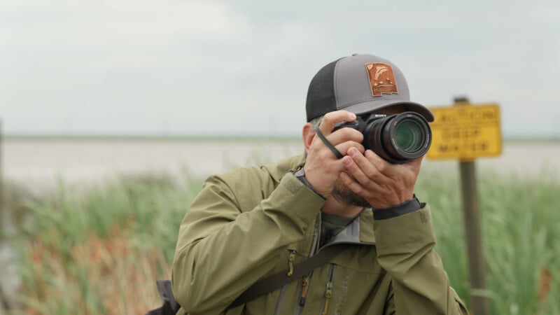 A person in a green jacket and cap is taking a photo with a camera outdoors near tall grass. A yellow sign is visible in the blurry background.