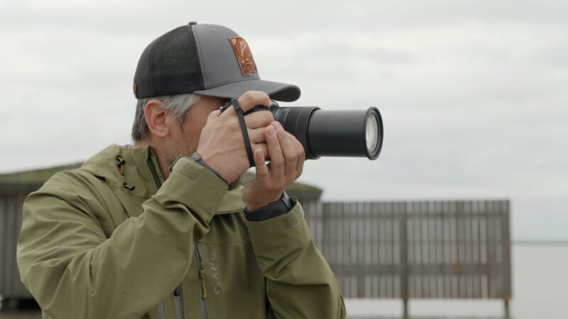 A person wearing a green jacket and dark cap is taking a photo with a large camera outdoors, with a wooden fence and cloudy sky in the background.