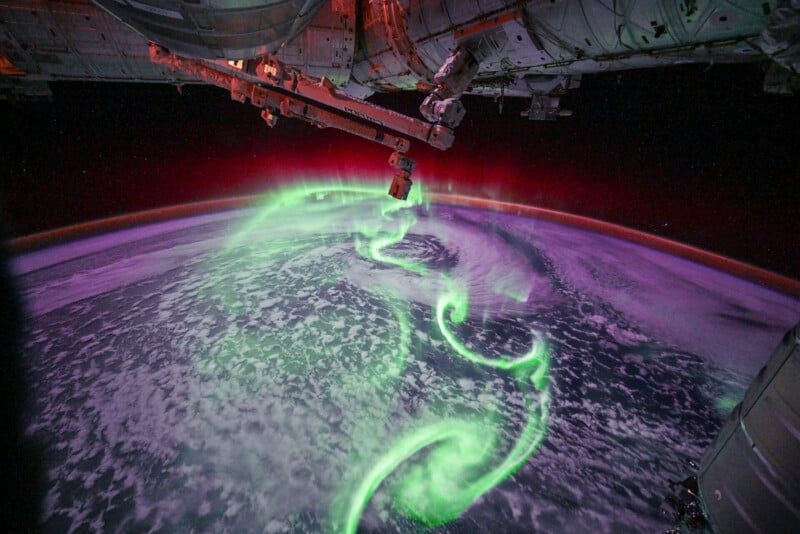 The image shows a vivid green aurora swirling over Earth's curved horizon, viewed from the International Space Station with part of the station’s structure visible at the top.