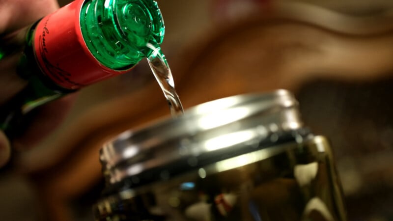 A close-up of liquid being poured from a green bottle with a red label into a glass jar. The background is blurred, focusing attention on the pouring action.