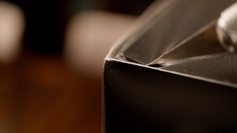 Close-up of a shiny, sharp-edged metallic object, possibly the corner of a box or a geometric sculpture, with blurred shapes in the background.