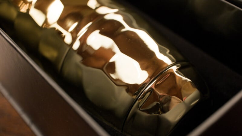 A close-up of a shiny, metallic gold cocktail shaker resting in a black box, reflecting surrounding light and colors.