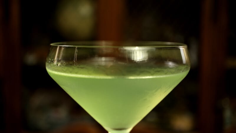 A close-up of a coupe glass filled with a bright green cocktail, with condensation on the glass and a blurred dark background.