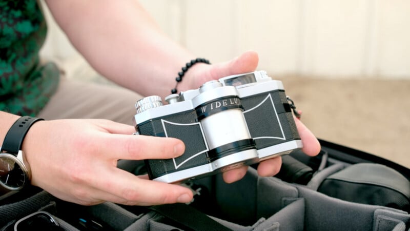 A person holding a vintage Widelux panoramic film camera above an open camera bag, with part of their watch and bracelet visible.