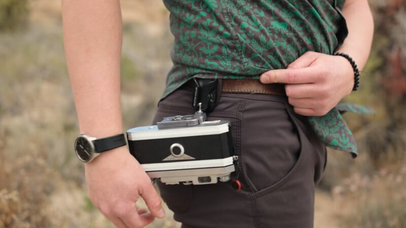 A person wearing a green patterned shirt and dark pants attaches a vintage camera to their belt using a clip. They have a watch on one wrist and a black beaded bracelet on the other. The background is outdoors and blurry.
