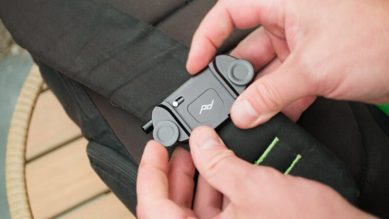 A person’s hands are attaching a black Peak Design clip to the strap of a black backpack. The clip has a logo and is secured using two round knobs. The background shows a wooden surface slightly out of focus.