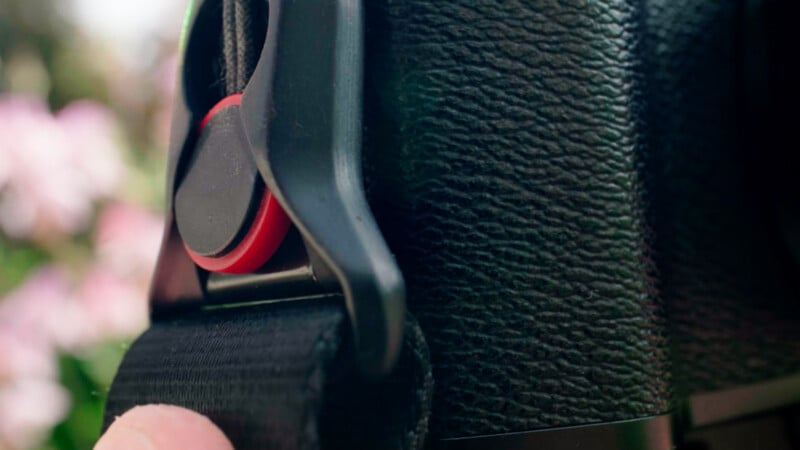 Close-up of a black camera strap attachment with a red accent, secured to the textured side of a camera body. A person's finger is visible holding part of the strap in the lower left corner.