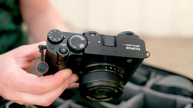 A person holds a Fujifilm GFX 100S camera above a cushioned camera case, showing the camera’s top controls, lens, and dials in detail.