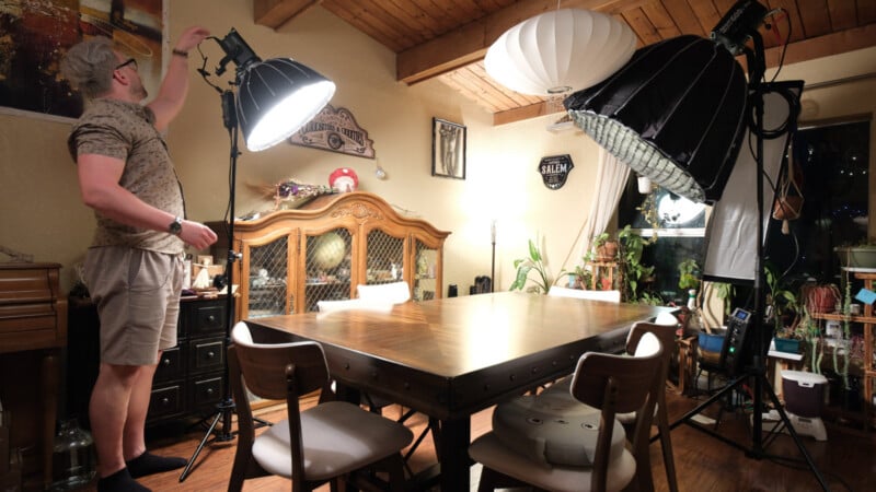 A person adjusts a lighting fixture in a cozy dining room with wooden ceiling, a set dining table, chairs, and plants. Large studio lights and photography equipment are set up around the room.