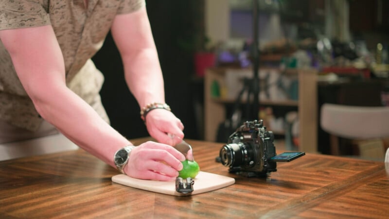 A person slices a lime on a wooden cutting board, with a digital camera and a small microphone placed nearby on the table.
