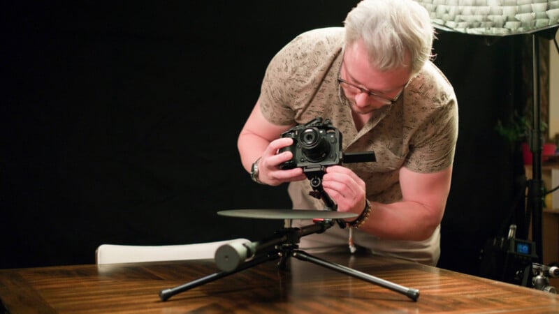 A person with gray hair and glasses adjusts a camera on a tripod, focusing on a round object on a table, with a dark background and studio lighting visible.
