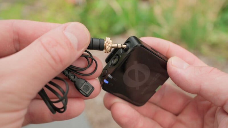 A person plugs a 3.5mm audio cable with a gold ring into a small black electronic device, possibly a portable audio recorder or wireless transmitter, outdoors with greenery blurred in the background.