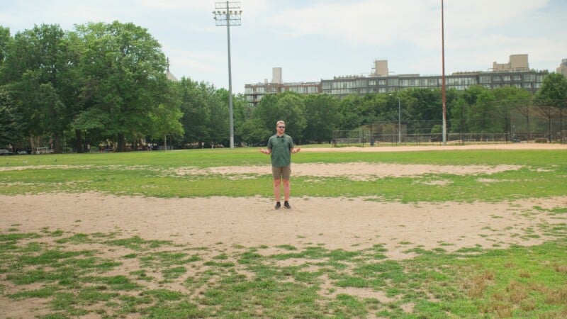 A person stands alone on a baseball field with grass and dirt patches. Trees and a building are in the background under a partly cloudy sky.