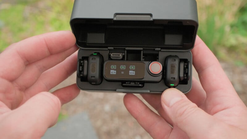 Two hands hold an open black charging case containing a compact wireless microphone system with two microphones and a receiver inside.