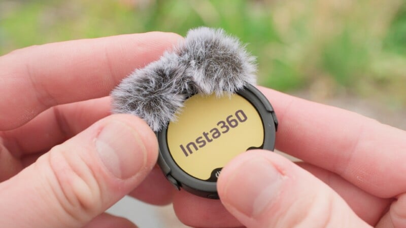 A person holds a small, round Insta360 device with a yellow face and fluffy gray windshields on top, outdoors with green foliage blurred in the background.