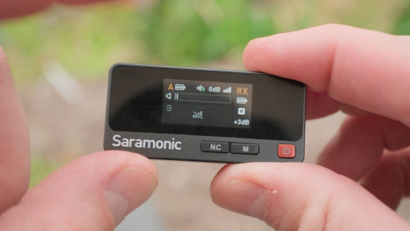 A person holds a small Saramonic audio device displaying input levels and settings on its screen. The device has a power button and controls labeled NC and M.