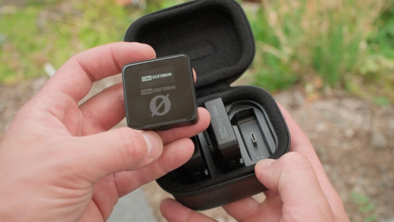 A person holds a small wireless microphone labeled "Wireless Pro" above an open black carrying case containing matching audio equipment. Greenery is blurred in the background.