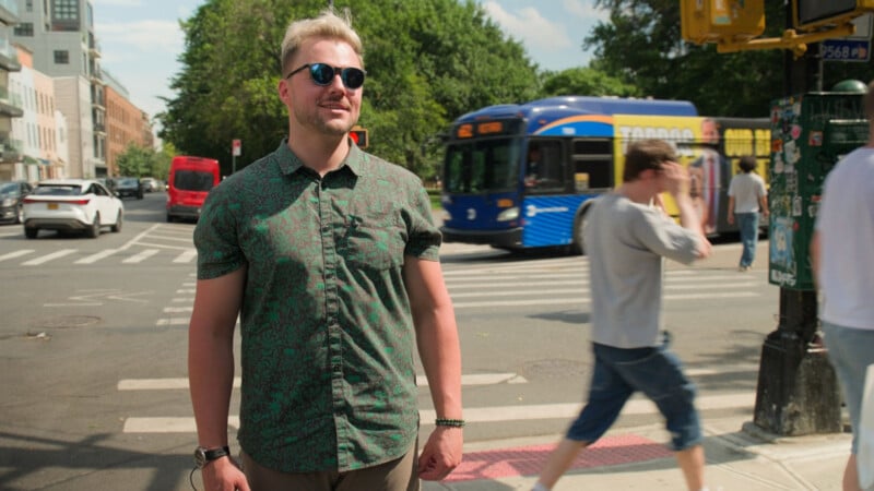 A man in sunglasses and a patterned shirt stands on a sunny city street corner. A blue bus, pedestrians, and cars are in the background.