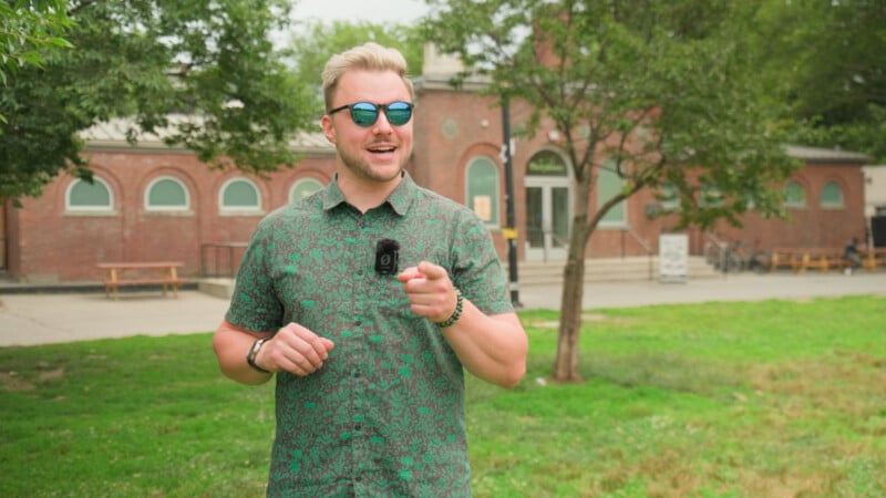 A man wearing sunglasses and a green patterned shirt smiles and points forward while standing on grass, with trees and a brick building in the background.