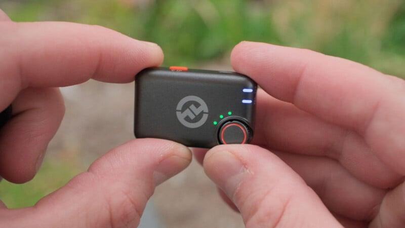 A person holds a small black electronic device with three blue LED lights, two green lights, a circular dial, and a logo on the front, using both hands.