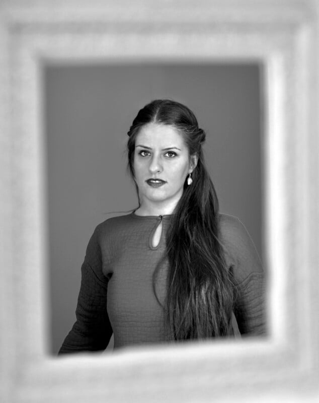 Black-and-white photo of a woman with long dark hair, wearing a textured dress with a keyhole neckline, looking at the camera through a blurred picture frame in the foreground.