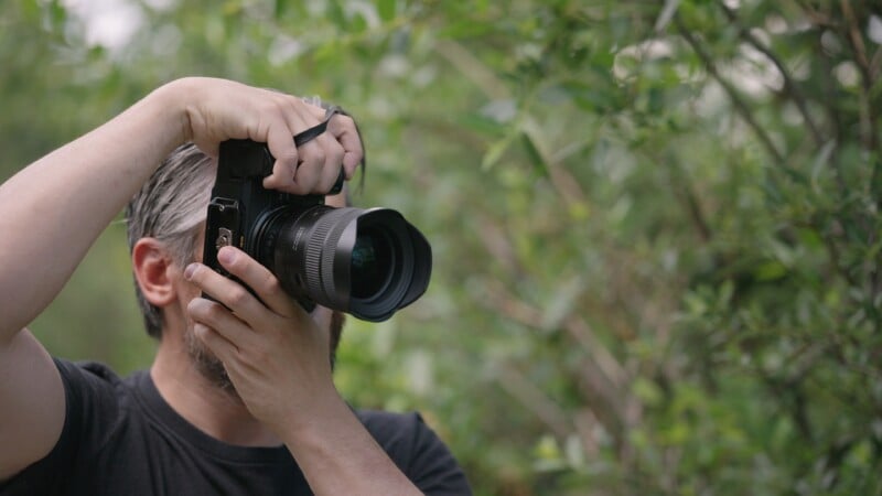 A person with gray hair and a beard is outdoors, holding a large camera up to their face, preparing to take a photo amid green foliage.