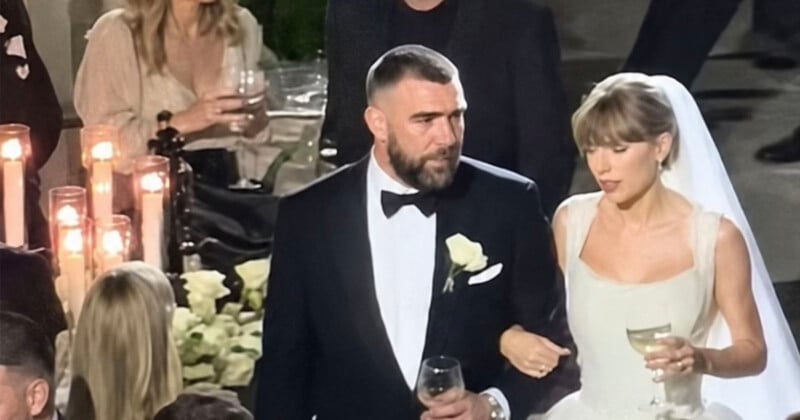 A bride in a white dress and veil walks arm-in-arm with a man in a black tuxedo, both holding wine glasses at a candlelit wedding reception with seated guests in the background.