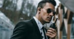 A man in a suit wearing sunglasses and an earpiece holds a walkie-talkie, appearing alert and focused, possibly acting as security or a bodyguard. Trees and modern buildings are visible in the background.