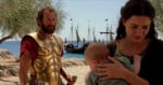 A man in golden armor stands near a woman holding a baby, with ships anchored by the shore and olive trees in the background.
