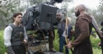 Actors in medieval costumes stand near a large film camera outdoors on a grassy set. Crew members adjust equipment while one actor holds a bow and another has arrows on his back.