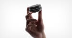 A hand holds a small camera lens delicately between the thumb and index finger against a plain white background.