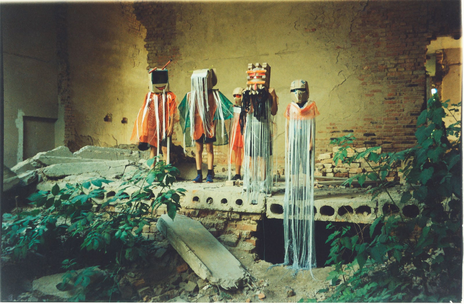 Five children wearing elaborate, handmade cardboard masks and long, fringed capes stand in a line amidst the ruins of a brick building.