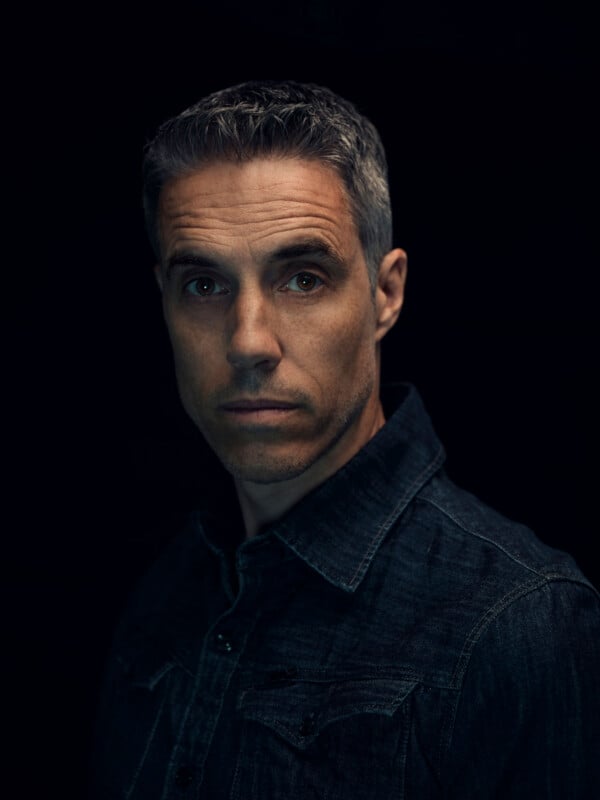 A man with short gray hair wearing a dark denim shirt looks seriously at the camera against a dark background.