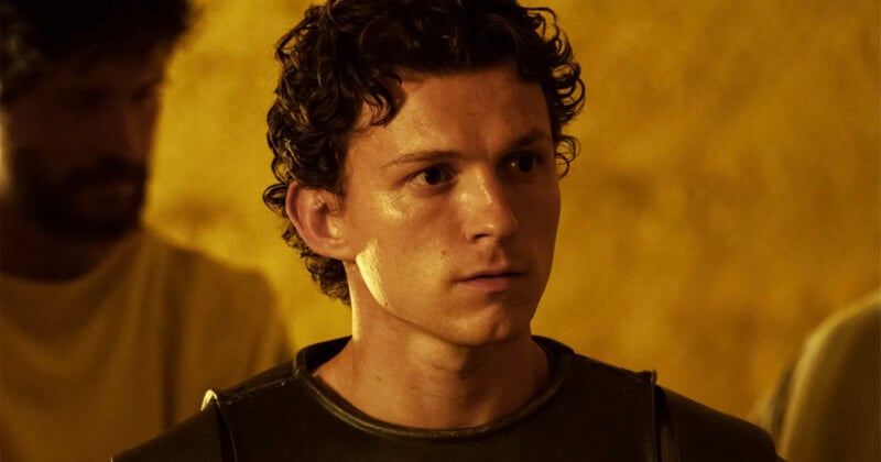 A young man with curly brown hair wearing a dark shirt and armor looks ahead seriously; the background is yellow with a blurred figure behind him.