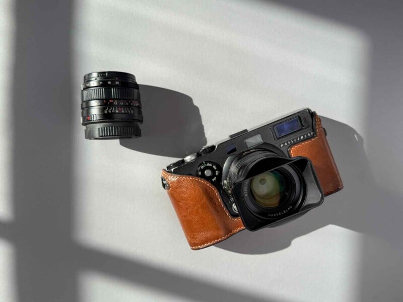 A camera with a brown leather case and a detached lens is placed on a light gray surface with shadows from a window falling across them.