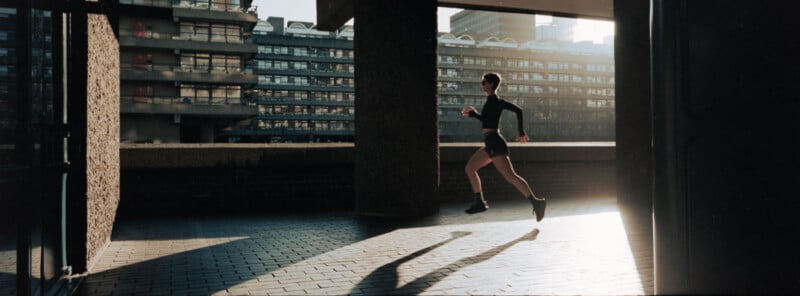 A person in athletic wear runs through a shaded urban corridor, with sunlight casting long shadows and modern apartment buildings visible in the background.