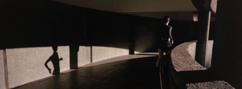 A person in dark clothing walks along a curved, shadowy concrete hallway, with their shadow cast on the wall.