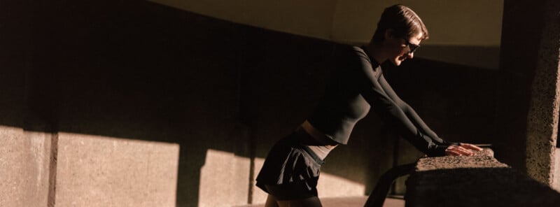 Person with short hair and sunglasses leans forward on a concrete railing in sunlight, wearing a black long-sleeve top and black skirt, with a shadowed background.