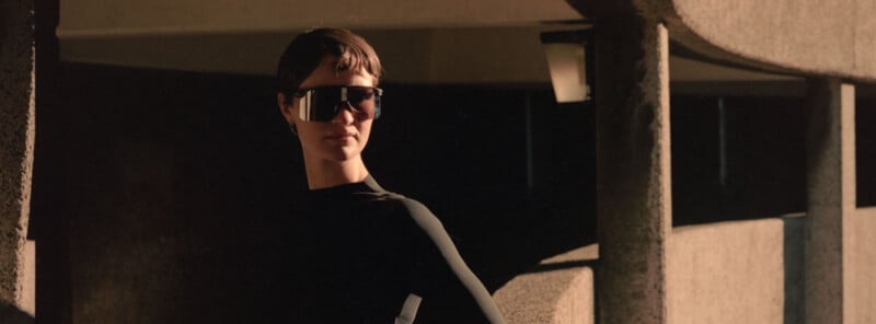 A person wearing large, futuristic sunglasses and a black top stands near concrete pillars in a sunlit, modern architectural setting.