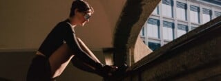 Person with short hair and sunglasses ties their shoe on a concrete ledge outdoors, with a building featuring blue window shades in the background.