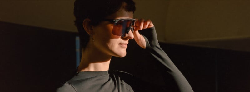 A person wearing a black long-sleeve top adjusts large, angular sunglasses while standing in soft, warm light against a dark background.