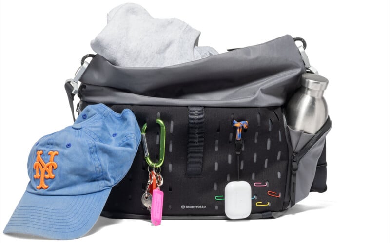 A black and gray shoulder bag with a water bottle in a side pocket, a sweatshirt inside, a blue New York Mets cap, colorful keychains, carabiners, AirPods case, and paperclips attached to the front.