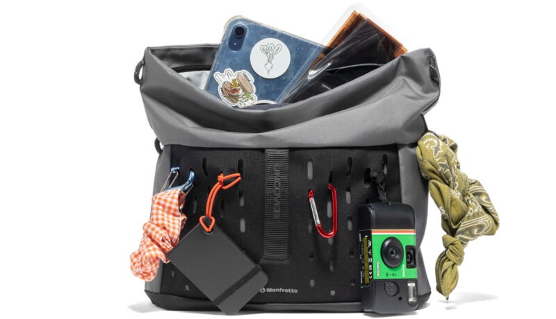 A gray and black camera bag filled with various items, including a tablet with stickers, a notebook, a bandana, a disposable camera, portable hard drive, and carabiners with keychains attached outside the bag.
