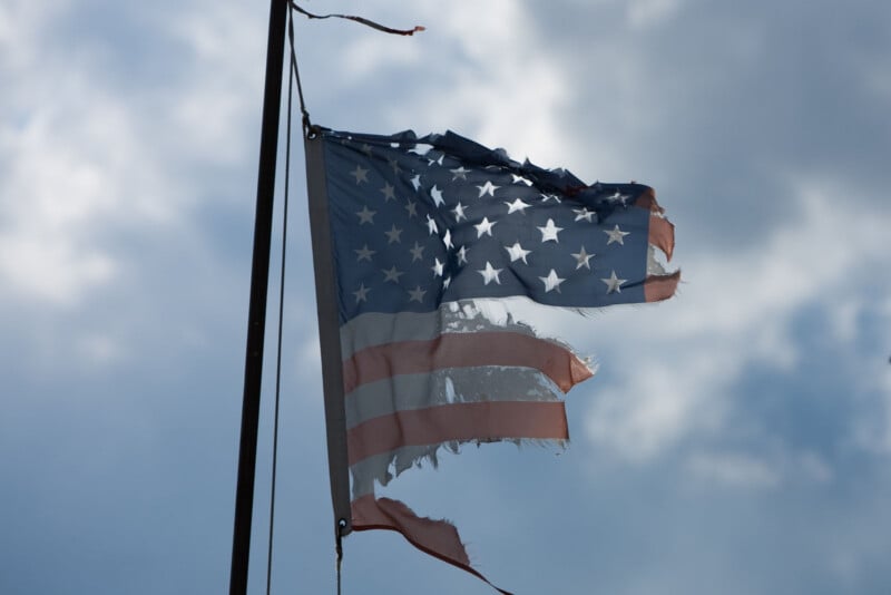A tattered and faded American flag with frayed edges blows in the wind against a cloudy sky.