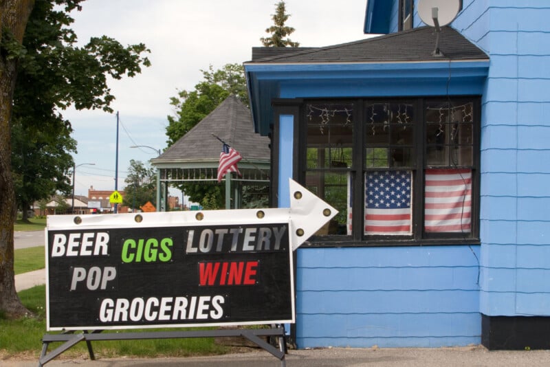 A large sign in front of a blue building advertises beer, cigs, lottery, wine, pop, and groceries. An arrow points to the entrance. An American flag hangs in the window, and another flies outside.