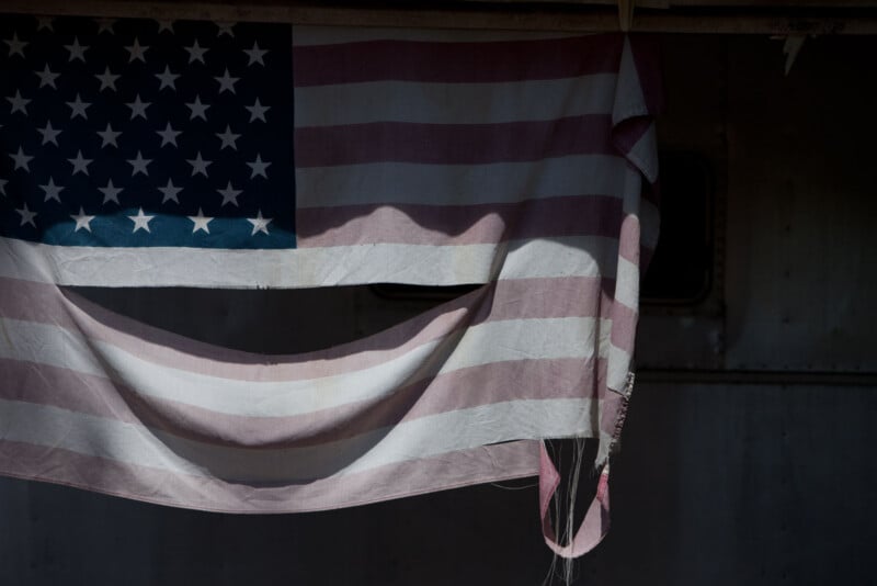 A faded and tattered American flag hangs, the bottom edge frayed and curling, with shadows cast across it. The background appears dark and metallic, suggesting an industrial or weathered setting.