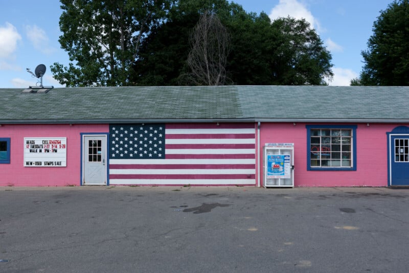 A pink building with a mural of the American flag painted on its front wall, a window, a vending machine, and a sign with business hours. There are trees and a blue sky in the background.