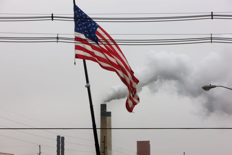 A tattered American flag hangs on a pole in the foreground, with industrial smokestacks in the background emitting plumes of smoke into a gray sky, intersected by several power lines.